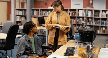 Estudante em cadeira de rodas ao lado de professora na biblioteca.