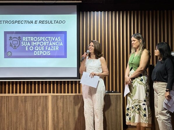 Mulher em palco, falando no microfone, ao lado de outra mulher.