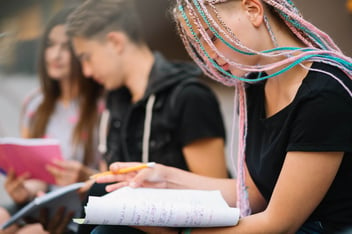 Menina com cabelos coloridos escrevendo, ao lado de um garoto e outra garota