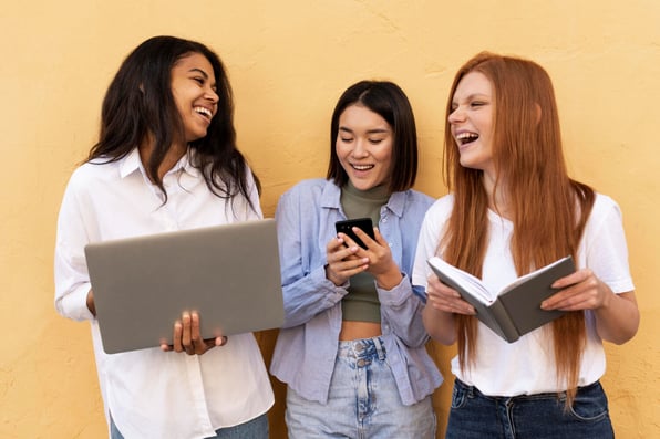 Três mulheres conversando e rindo entre si enquanto seguram dispositivos eletrônicos.