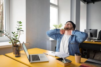 homem sentado a mesa, em frente ao seu notebook, se espreguiçando.