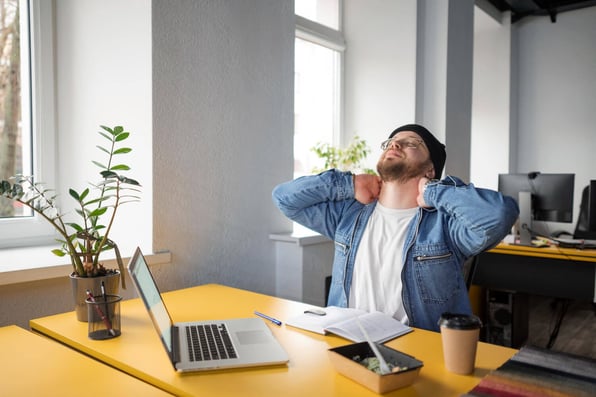 homem sentado a mesa, em frente ao seu notebook, se espreguiçando.