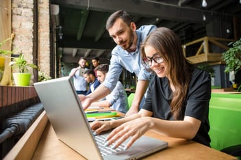 Menina escrevendo no notebook enquanto recebe orientações de um homem.