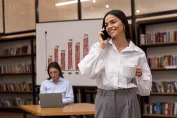 Mulher ao telefone, em frente a outra mulher que se encontra trabalhando sentada a mesa, com seu notebook e um quadro com um gráfico.