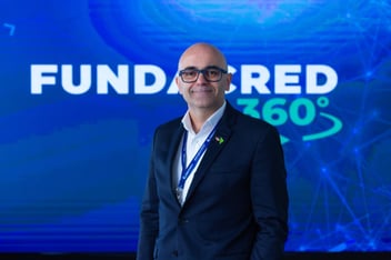 Homem branco, careca, de óculos, vestido com um terno, centralizado na imagem a frente de um painel escrito Fundacred 360