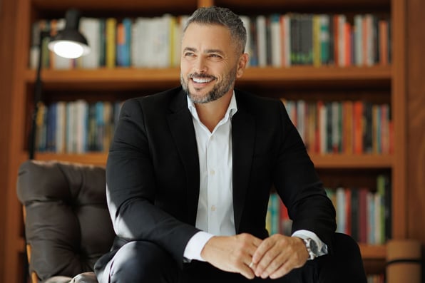 Homem branco, com cabelos e barba grisalhos, sorrindo, sentado a frente de uma estante de livros. Ele está usando um blazer preto e camisa branca.