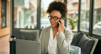 Mulher negra, com roupa social, sentada a mesa, mexendo em notebook, enquanto fala ao telefone.