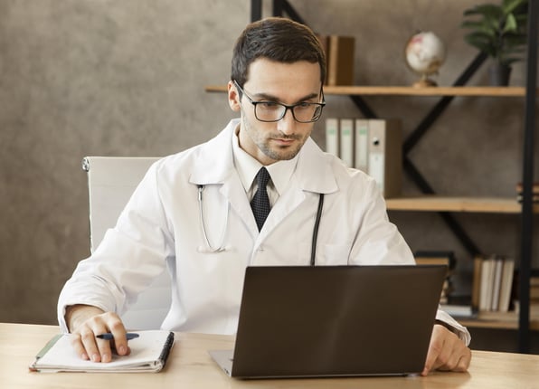 Médico sentado a mesa, em frente a notebook.