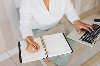 Pessoa sentada em uma mesa mexendo no notebook e escrevendo em um caderno.