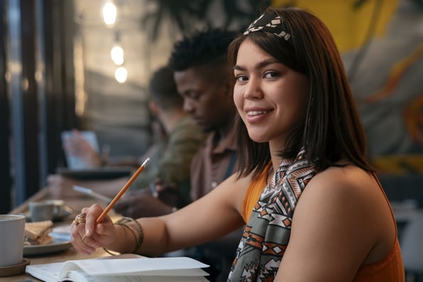 Jovem mulher, com traços indígenas, sentada a mesa, escrevendo em um caderno.