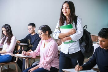 Estudante em sala de aula carregando livros e cercada por colegas