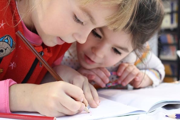 Uma menina escrevendo em uma folha de papel enquanto a outra observa 